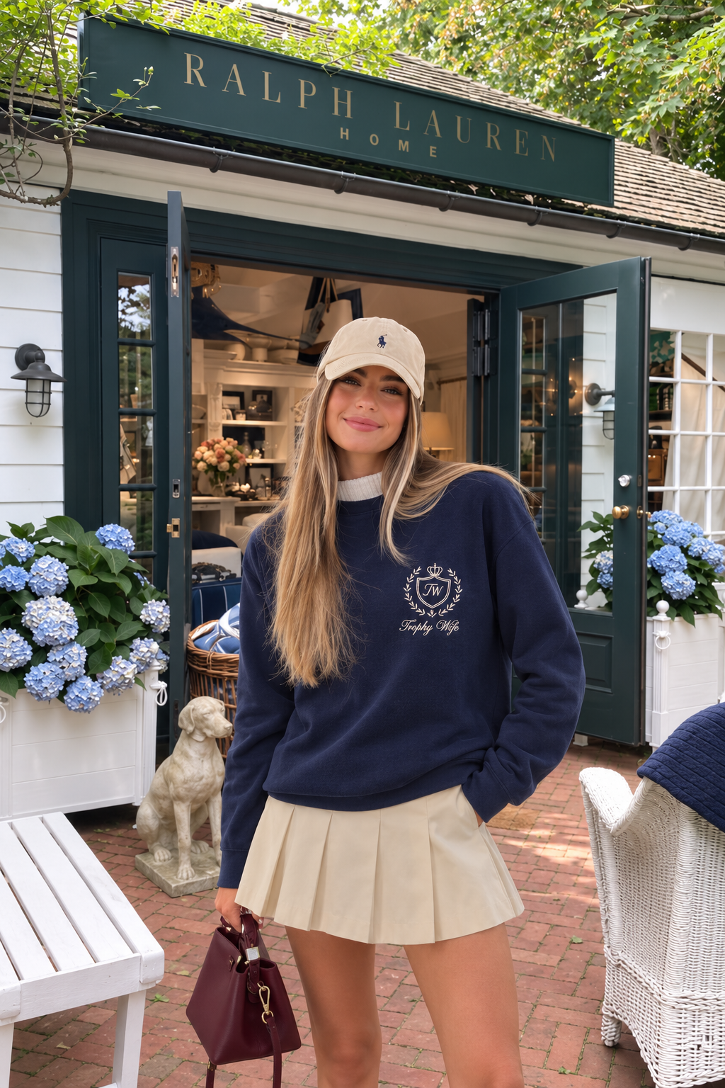 A woman stands in front of a Ralph Lauren Home storefront wearing a navy crewneck sweatshirt with a white crest graphic, a beige pleated mini skirt, and a beige baseball cap. She is holding a burgundy handbag.